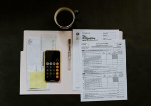 foto de uma pasta aberta com papéis em cima, um celular com a calculadora aberta e um café acima da imagem para simular a realização de um balanço patrimonial